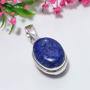 May include: An oval-shaped lapis lazuli pendant set in a polished silver frame. The deep blue stone features natural gold-colored pyrite flecks throughout its surface. A silver bail is attached to the top of the setting for hanging on a chain.