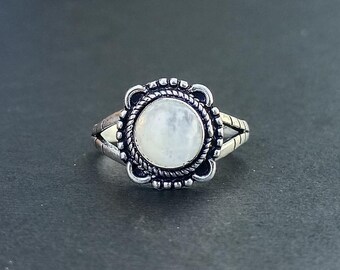May include: A silver-toned ring with a large, round, pale gemstone center. The gemstone is surrounded by a decorative border with a rope-like texture. The ring has a split band design with decorative accents.