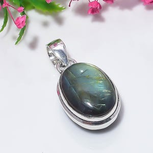 May include: An oval-shaped pendant featuring a polished, dark green and grey labradorite gemstone set in a smooth, shiny silver metal frame. The pendant includes a silver bail at the top for attachment, showcasing natural iridescent inclusions.