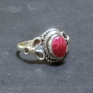 Può includere: Un anello in argento con una pietra rossa di forma ovale al centro, incastonata in un bordo di metallo intrecciato. La fascia presenta un design in metallo traforato e simmetrico su entrambi i lati, per un aspetto dettagliato.
