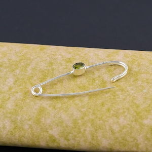 Może przedstawiać: Srebrna agrafka z okrągłym, jasnozielonym kamieniem szlachetnym osadzonym blisko środka. Agrafka jest otwarta, z ostrym końcem skierowanym w dół. Kamień szlachetny ma żywy, oliwkowy kolor.