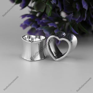 Puede incluir: Dos expansores de oreja plateados en forma de corazón. Uno es un rectángulo sólido y redondeado con una parte superior en forma de corazón. El otro es un túnel en forma de corazón. Flores moradas en el fondo.