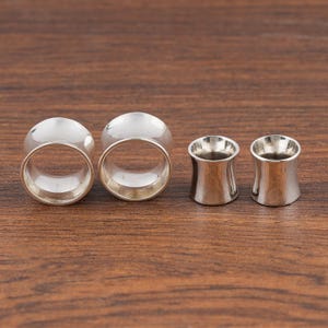 Op de afbeelding: Vier zilverkleurige oor stretchers op een houten oppervlak. Twee zijn grote, afgeronde tunnels en twee zijn kleinere, uitlopende plugs. De stretchers zijn gemaakt van een glanzend, metalen materiaal. De achtergrond is een warme, bruine houtnerf.