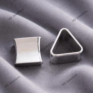 May include: Two silver-colored plugs, one square and one triangular, sit on a gray fabric. The square plug has a curved design, while the triangle plug has a simple, geometric shape. The plugs are likely for ear piercings.