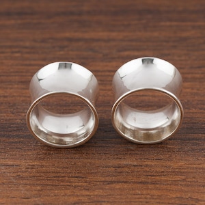 May include: Two polished silver tunnels, designed for stretched ear piercings, are displayed against a warm, brown wooden surface. The tunnels have a flared design, with a wider front and back, and a smooth, reflective surface.