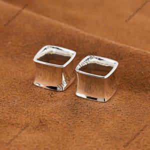 Op de afbeelding: Twee gepolijste zilveren vierkante oordopjes. De oordopjes hebben een licht gebogen ontwerp en worden gepresenteerd op een bruin oppervlak. De sieraden zijn reflecterend en lijken van metaal te zijn.