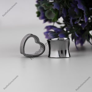 Puede incluir: Dos piezas de joyería corporal de metal gris oscuro. Una es un tapón en forma de corazón y la otra es un tapón cilíndrico. Las joyas se exhiben sobre una superficie blanca con un arreglo floral morado borroso en el fondo.
