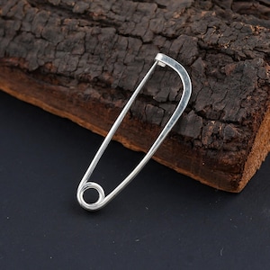 Może przedstawiać: Srebrna agrafka z zaokrąglonym zapięciem i spiczastym końcem. Szpilka wykonana jest z błyszczącego metalu i umieszczona na ciemnym tle z kawałkiem teksturowanego drewna.