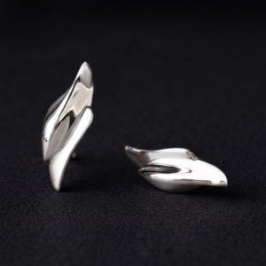Op de afbeelding: Een paar oorbellen van gepolijst zilver met een gladde, gebogen, bladvormige vorm. Het metaal heeft een reflecterende, hoogglanzende afwerking met vloeiende, organische contouren die toelopen in een punt voor een strak en modern uiterlijk.
