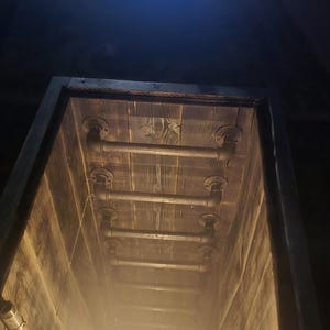 Puede incluir: Estructura de madera con tuberías y accesorios metálicos, iluminada por una luz cálida en la parte inferior. El interior está revestido con tablones de madera, creando un efecto de túnel. Las tuberías están dispuestas horizontalmente en el techo.