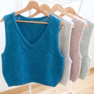 Peut inclure: Présentation de quatre gilets tricotés de couleurs différentes : bleu sarcelle, gris, rose et gris clair. Chaque gilet a une encolure en V et est suspendu à un cintre en bois. Les gilets sont exposés sur un portant blanc.