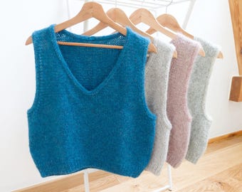 May include: A display of four knitted vests in various colors: teal, gray, pink, and light gray. Each vest has a V-neck design and is hanging on a wooden hanger. The vests are displayed on a white rack against a white wall.