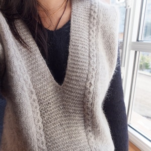 May include: A light gray knitted vest with a deep V-neck. The vest has a textured pattern along the sides and is worn over a dark blue sweater. The image is taken indoors, near a window.