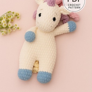 May include: A handmade crochet unicorn plushie in cream, with blue hooves and accents, and a purple mane and horn. The image includes a "PDF Crochet Pattern" graphic. The unicorn is posed on a pink background.