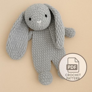 Puede incluir: Un juguete de conejo de ganchillo gris suave con largas orejas caídas y un cuerpo redondeado. El conejo tiene ojos negros y una nariz blanca. Un icono de patrón de ganchillo PDF está en la esquina inferior derecha.
