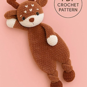 May include: A brown crochet deer plushie with white accents on the face, antlers, and feet. The deer has a white belly and blue eyes. The text "PDF CROCHET PATTERN" is in a circle in the upper right corner. The deer is lying on a pink background.