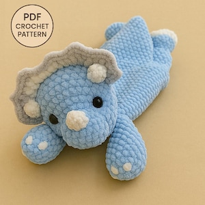 May include: A light blue crocheted Triceratops dinosaur toy with gray and white accents. The dinosaur has black eyes and white spots on its feet. The image includes a circular graphic that says "PDF CROCHET PATTERN".