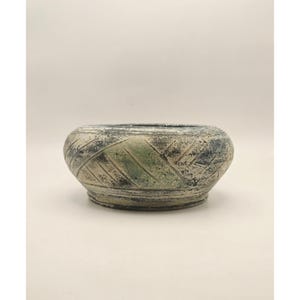 May include: A low, rounded ceramic bowl featuring a textured surface with a distressed finish. The exterior displays a blend of muted green, charcoal grey, and off-white tones, accented by a series of diagonal incised lines around the body.