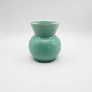 May include: A small, smooth ceramic vase with a rounded, bulbous body and a flared, wide opening at the top. The entire exterior surface is finished in a solid, matte mint green glaze, providing a consistent colour throughout the piece.