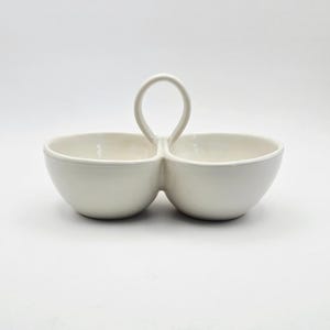 May include: A white ceramic serving dish with two connected bowls and a handle. The bowls are round and the handle is a loop shape. The dish is on a white background.