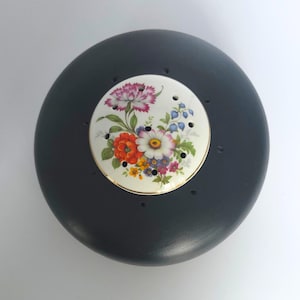 Puede incluir: Un tope de flores redondo de madera gris oscuro con una inserción de cerámica blanca. La inserción presenta un diseño floral con flores rojas, naranjas, moradas y blancas. El tope tiene varios agujeros pequeños.