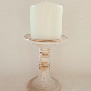 May include: A cream-coloured pillar candle on a ceramic candle holder. The holder has a pedestal base and a tiered stem. The candle is unscented and has a single wick. The candle holder is a neutral beige colour.