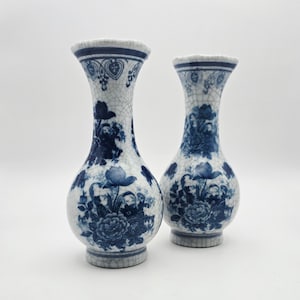 May include: Two ceramic vases featuring a white base with a blue floral pattern and a distinct crackle glaze finish. Each vase has a bulbous body, a slender neck, and a flared rim, decorated with detailed botanical illustrations in deep blue.