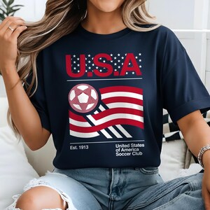 May include: Navy blue t-shirt with a graphic featuring the text "U.S.A." in red, a soccer ball, and a stylized American flag. The shirt also includes the text "Est. 1913" and "United States of America Soccer Club."