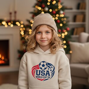 May include: A young person wearing a beige hoodie and a matching beanie. The hoodie features a graphic with a soccer ball, the text "USA", and "WORLD CUP '26" in red and blue. The background includes a Christmas tree and a fireplace.