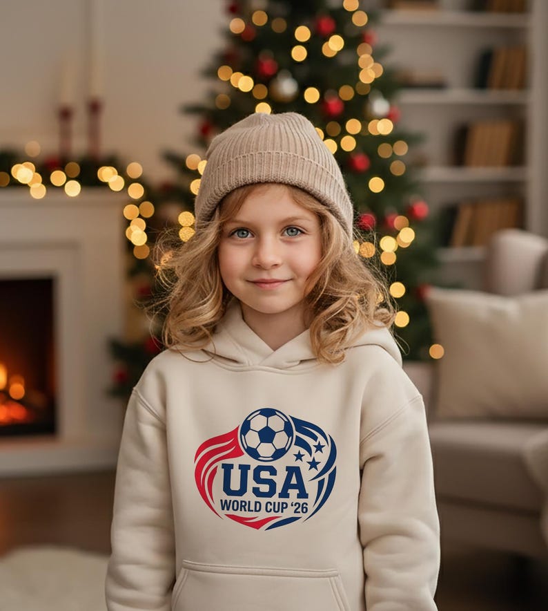 May include: A young person wearing a beige hoodie and a matching beanie. The hoodie features a graphic with a soccer ball, the text "USA", and "WORLD CUP '26" in red and blue. The background includes a Christmas tree and a fireplace.