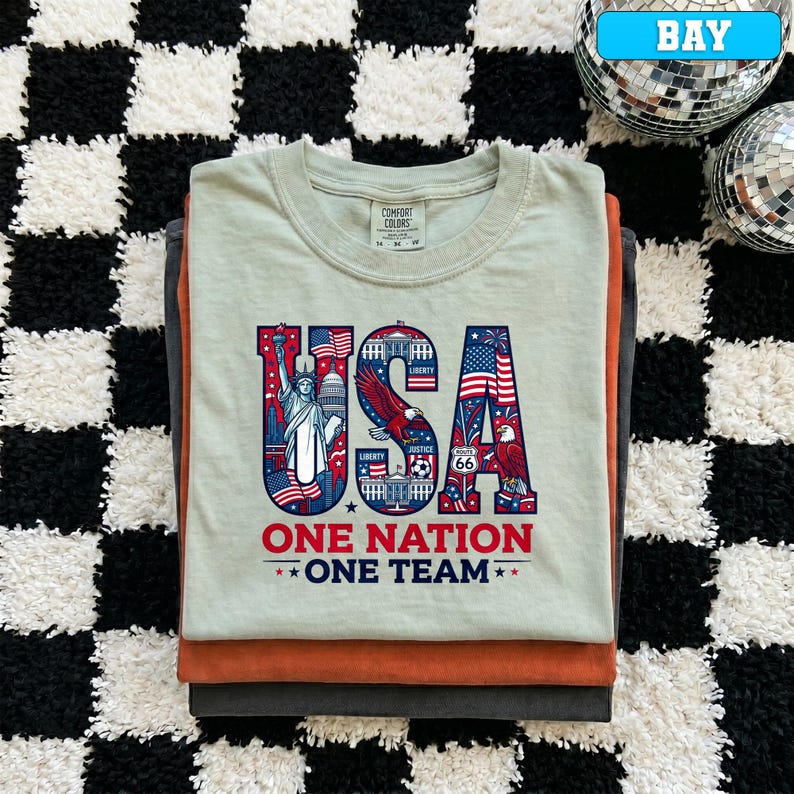 May include: A stack of folded t-shirts. The top shirt is light green with a graphic that says "USA ONE NATION ONE TEAM" in red, white, and blue. The graphic includes the Statue of Liberty, an eagle, and the American flag. The other shirts are orange and gray.