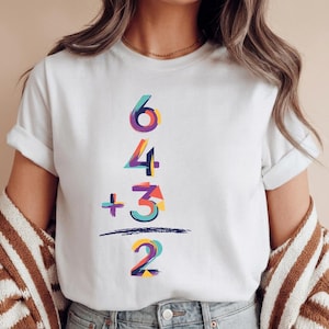 Könnte beinhalten: Weißes T-Shirt mit einem farbenfrohen mathematischen Gleichungsdesign. Die Gleichung lautet "6 4 + 3 2", wobei jede Zahl eine andere Farbe hat. Die Zahlen sind vertikal gestapelt, getrennt durch einen horizontalen Strich.