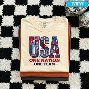 May include: A stack of folded t-shirts in ivory, rust, and charcoal colors. The top shirt features a graphic design with the word "USA" in red, white, and blue, along with the text "ONE NATION ONE TEAM".