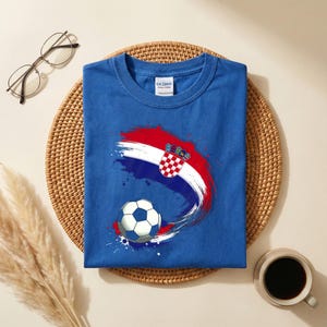 May include: A blue t-shirt featuring a Croatian flag design with a soccer ball graphic. The design incorporates red, white, and blue brushstrokes and the Croatian coat of arms. The shirt is made of cotton.