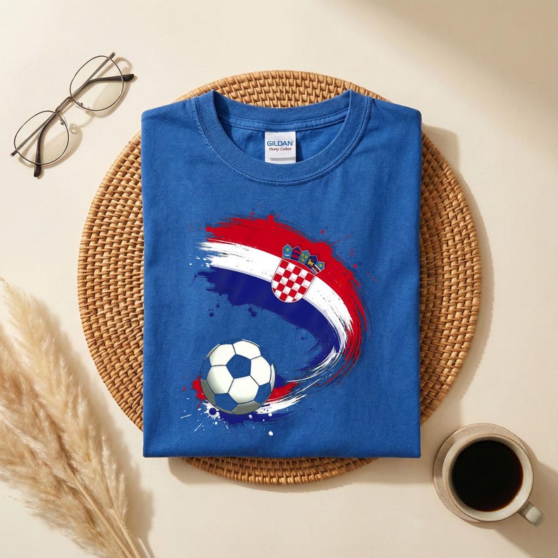 May include: A blue t-shirt featuring a Croatian flag design with a soccer ball graphic. The design incorporates red, white, and blue brushstrokes and the Croatian coat of arms. The shirt is made of cotton.
