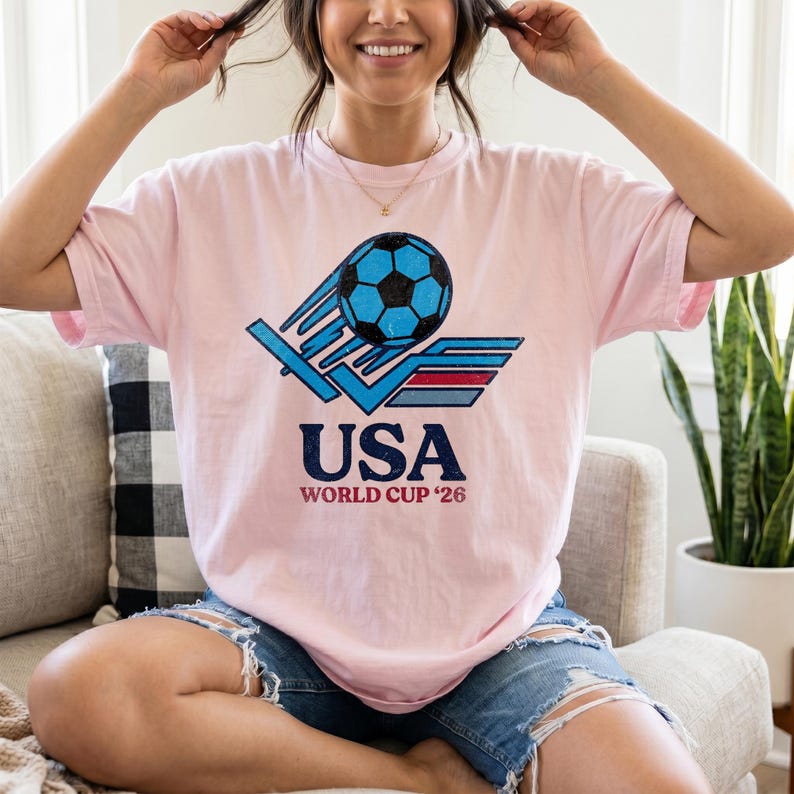 Puede incluir: Camiseta rosa claro con un dise&ntilde;o gr&aacute;fico de bal&oacute;n de f&uacute;tbol azul y el texto "USA World Cup '26" en azul marino. El dise&ntilde;o incluye un efecto de estela. Cuello redondo y mangas cortas.