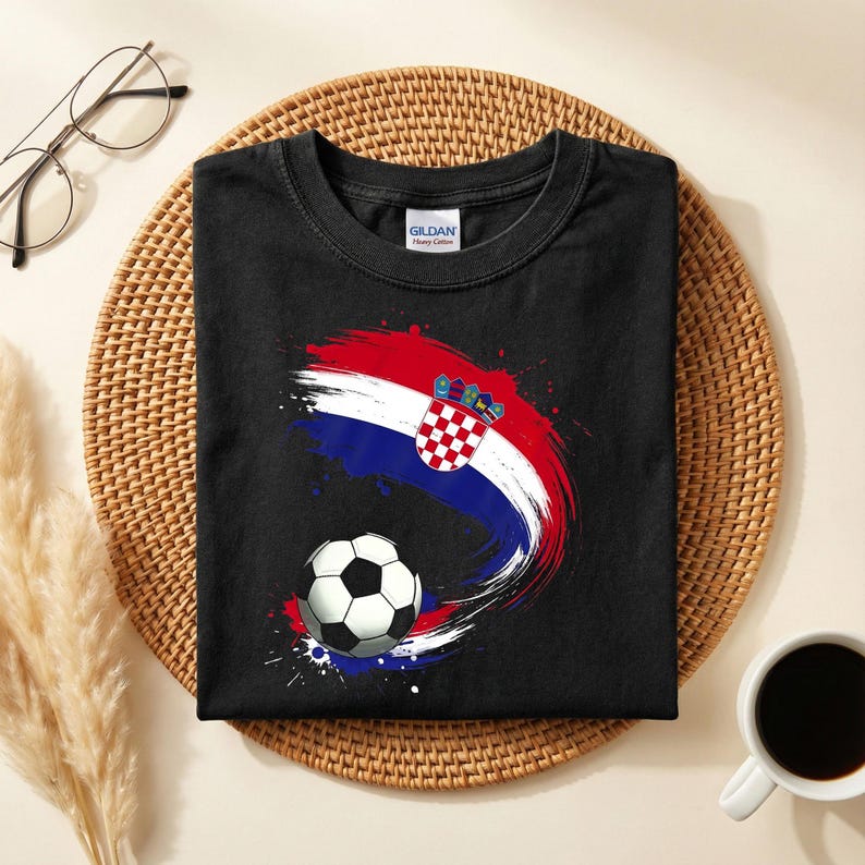 May include: Black t-shirt featuring a soccer ball and a design in the colors of the Croatian flag: red, white, and blue. The design has a brushstroke effect. The shirt is folded on a woven mat.