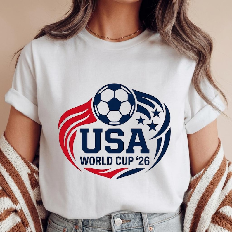 May include: White t-shirt featuring a soccer ball graphic with the text "USA WORLD CUP '26". The design includes red and blue elements, with stars and curved lines. The shirt is a crew neck.