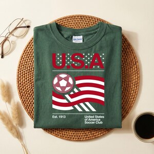 May include: Dark green t-shirt with a graphic featuring "U.S.A." in red letters with white stars, a soccer ball, and a stylized American flag. The shirt also includes the text "United States of America Soccer Club" and "Est. 1913".
