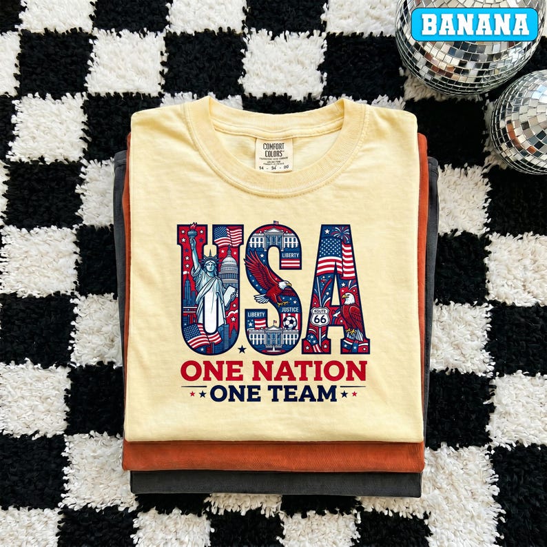 May include: A stack of three t-shirts. The top shirt is light yellow with the word "USA" in red, white, and blue, featuring the Statue of Liberty, an eagle, and flags. The text "ONE NATION ONE TEAM" is below. The other shirts are orange and gray.
