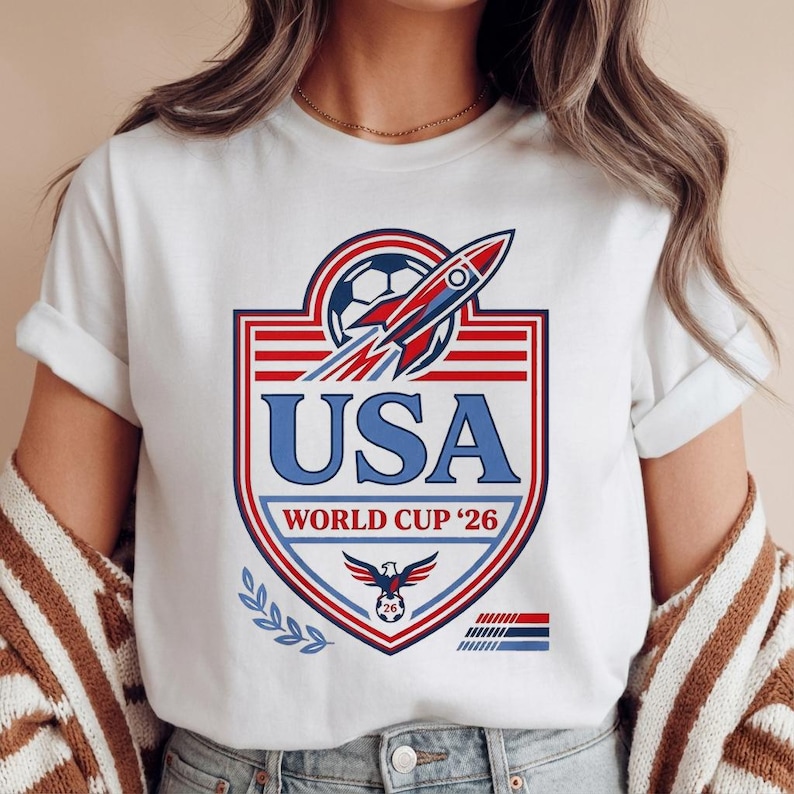 Peut inclure: T-shirt blanc avec un motif graphique pour la Coupe du Monde de la FIFA 2026 aux &Eacute;tats-Unis. Le motif comprend un ballon de football, une fus&eacute;e et le texte "USA" en bleu, avec des accents rouges et bleus. Un aigle patriotique est &eacute;galement pr&eacute;sent.