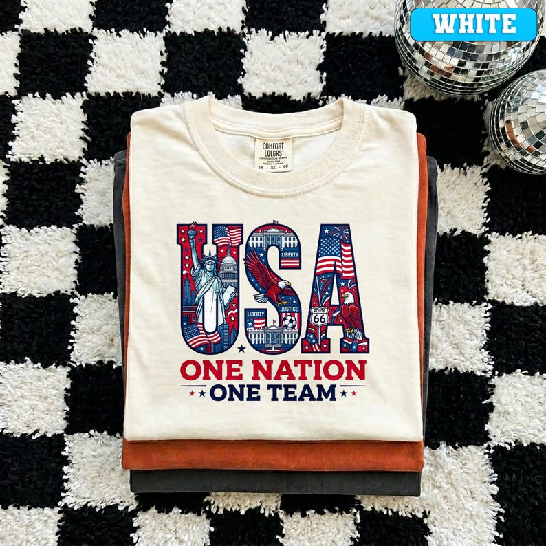 May include: Cream-colored t-shirt with a patriotic "USA" graphic featuring the Statue of Liberty, an eagle, and American flags. The text "ONE NATION ONE TEAM" is below the graphic. The shirt is folded on top of other shirts.