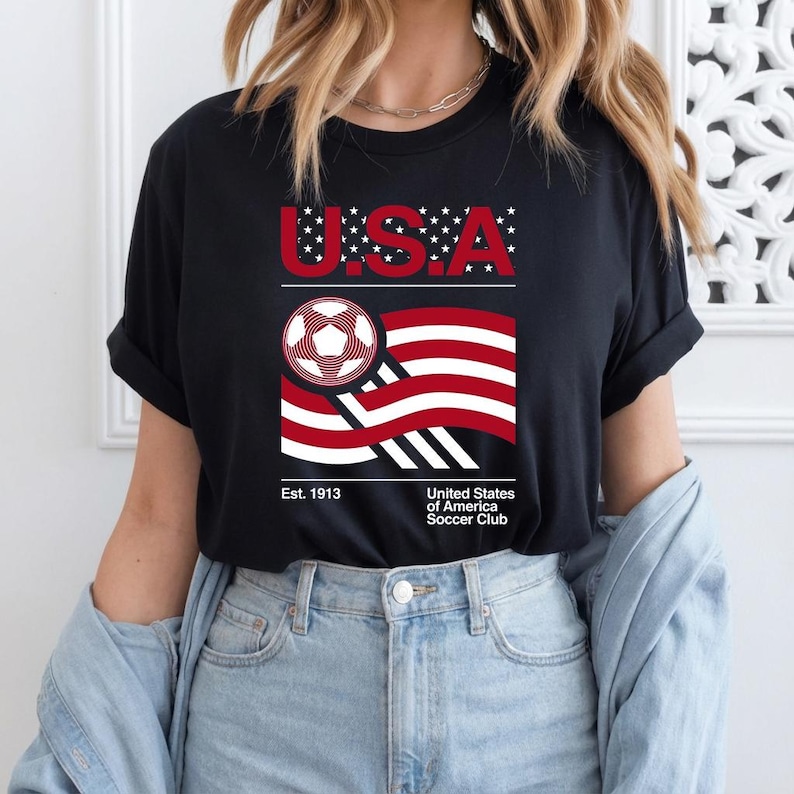 May include: Black t-shirt with a graphic design featuring the word "U.S.A." in red and white, a soccer ball, and a stylized American flag. The text "Est. 1913" and "United States of America Soccer Club" are also present.