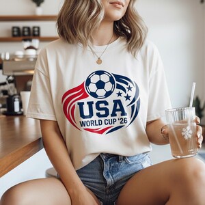 May include: Cream-colored t-shirt with a soccer ball graphic and the text "USA WORLD CUP '26" in red and blue. The shirt is worn with denim shorts. A clear glass with a straw is held in the hand.