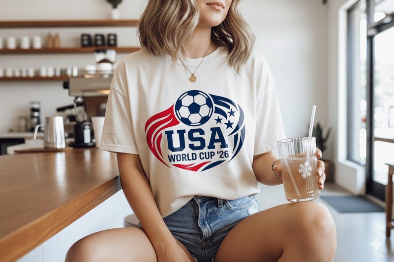 May include: Cream-colored t-shirt with a soccer ball graphic and the text "USA WORLD CUP '26" in red and blue. The shirt is worn with denim shorts. A clear glass with a straw is held in the hand.