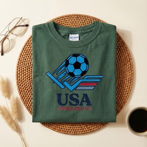 Puede incluir: Camiseta verde con un gr&aacute;fico de f&uacute;tbol retro. El dise&ntilde;o presenta un bal&oacute;n de f&uacute;tbol azul con l&iacute;neas de movimiento, la palabra "USA" y "WORLD CUP 76" en una fuente vintage. La camiseta est&aacute; doblada sobre una estera tejida.