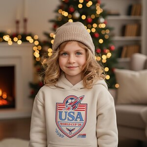 Peut inclure: Un sweat &agrave; capuche beige avec un motif graphique repr&eacute;sentant un ballon de football, une fus&eacute;e et le texte "USA WORLD CUP '26". Le sweat &agrave; capuche a une poche avant et une capuche &agrave; cordon. La personne porte un bonnet beige.