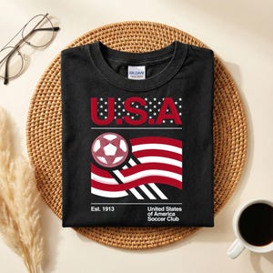 May include: Black t-shirt with a graphic design featuring "U.S.A." in red letters, a soccer ball, and a stylized American flag. The shirt includes the text "United States of America Soccer Club" and "Est. 1913".