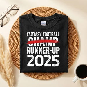 May include: Black t-shirt with white text that reads "FANTASY FOOTBALL CHAMP" with "CHAMP" crossed out in red, followed by "RUNNER-UP 2025". The shirt is folded on a woven mat, with a cup of coffee and glasses in the background.
