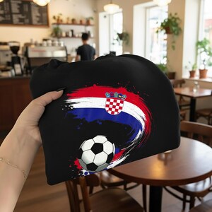May include: Black sweatshirt featuring a soccer ball and a design in the colors of the Croatian flag: red, white, and blue. The design includes the Croatian coat of arms. The sweatshirt is held up in front of a cafe setting.